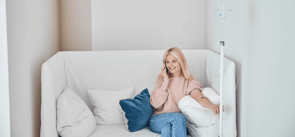 A woman sits on a white couch, talking on the phone while receiving IV therapy through her arm, surrounded by pillows.