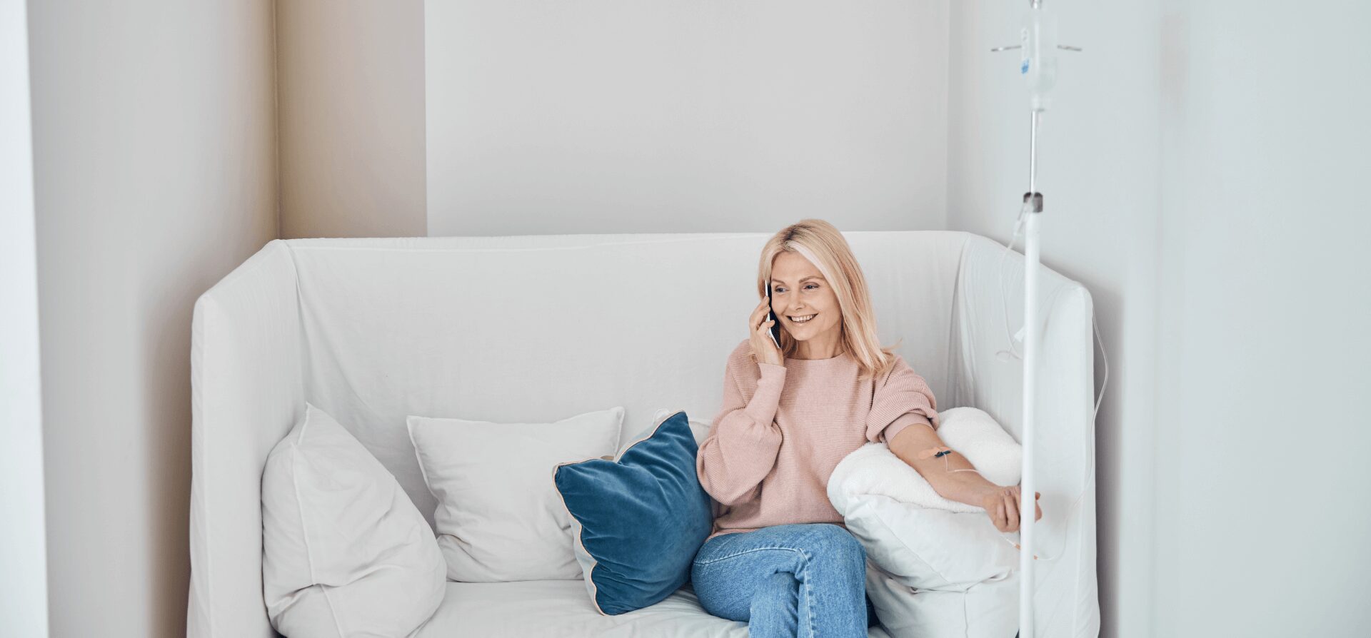 A woman sits on a white couch, talking on the phone while receiving IV therapy through her arm, surrounded by pillows.
