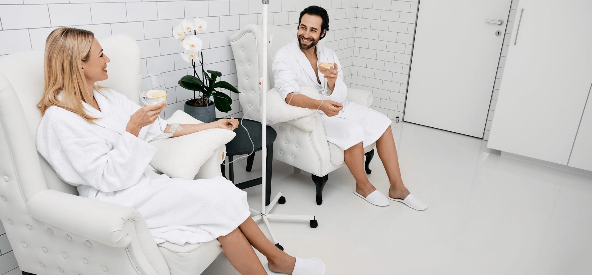 A woman and man in white robes sit in armchairs, smiling and holding drinks, with an IV stand between them in a modern, white-tiled room.