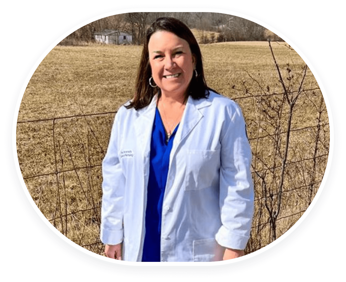 Woman in a white coat smiling outdoors in a rural setting.